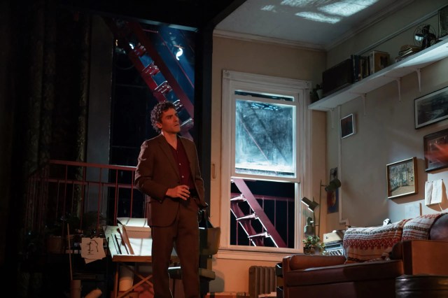 Oscar Isaac stands in front of a window and fire escape.