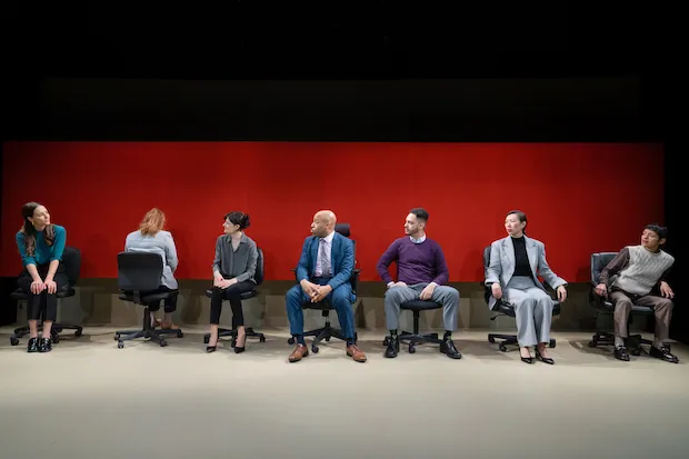 Actors sit an a row of swivel chairs against a red-orange backdrop. 