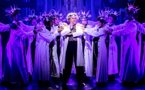 Roger Bart surrounded by ensemble members in lab coats.