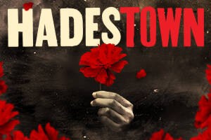A promotional poster for the musical Hadestown, the image has a close-up of a woman's hand holding a red rose against a black background
