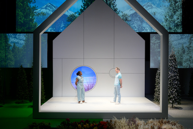 Jennifer Kidwell and Steven Rishard wear light blue outfits and stand facing each other, hands behind their backs, inside a white house-shaped structure with a circular blue and purple-lit window. Behind are screens projecting trees and mountains. Green grass is to the right and snow is to the left. 
