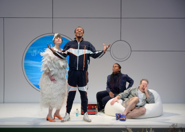 Brandon J. Pierce stands with his arms out wide looking joyously up into the sky. He is in a sweat suit in various shades of blue and an orange stripe. Sarah Gliko looks on with him standing just behind his right side, still in her white feathers, now in orange kitten heels. To Peirce’s left side, his parents sit on a bean bag chair, looking surprised. Jennifer Kidwell is in an all-navy business casual suit and Steven Rishard is in a light blue robe, underwear and slippers.