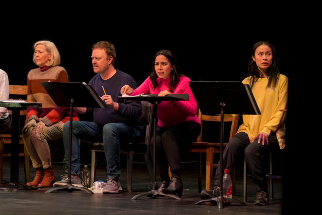 Four actors sit on stage in a straight line. In front of them, music stands holding binders.