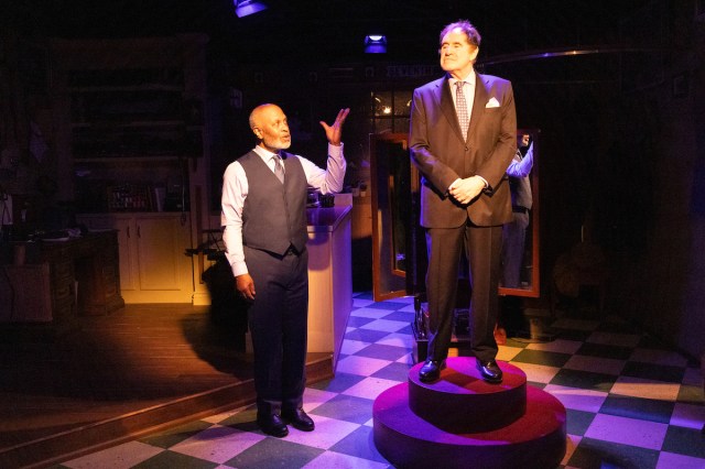 A man in a vest gestures toward a man in a suit standing on a pedestal. 