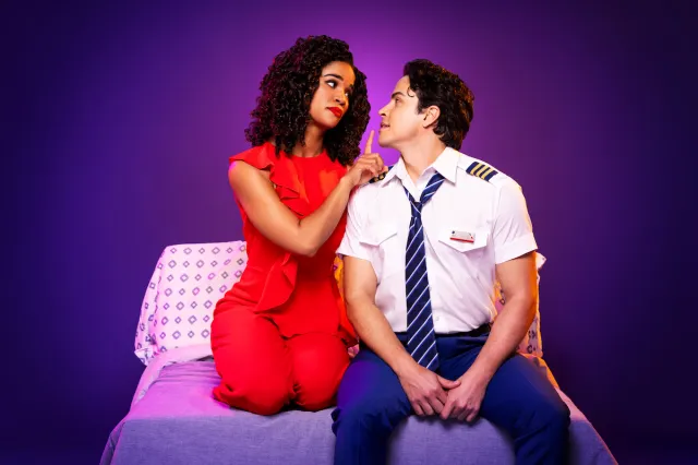 A woman holds her finger to the lips of a man in a flight uniform. Both are seated on a bed. 