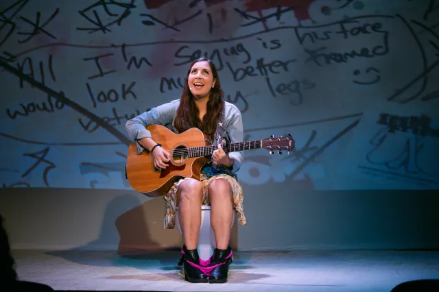 A woman sits on the toilet, strumming a guitar. 