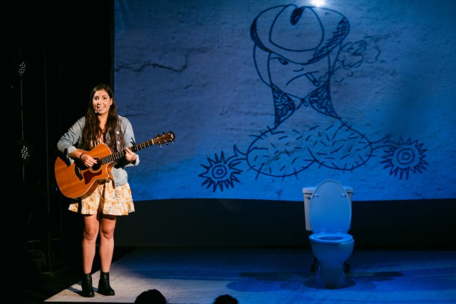 A woman plays her guitar in front of a large drawing of a penis cowboy. 
