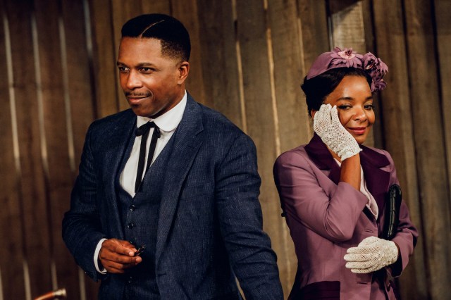 A man in a vintage blue three-piece suit looks away, while a woman in a vintage magenta suit holds a gloved hand to her face. 