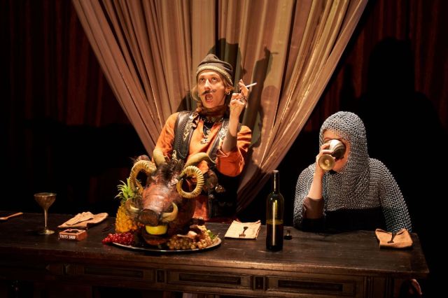 A woman dressed as a man smokes a cigarette behind an animal head on a platter. Another actor in chain mail drinks from a goblet. 