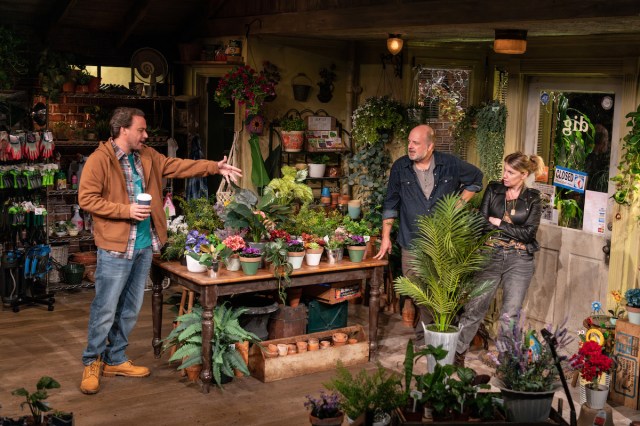 A man holds a paper cup of coffee and gestures over a table full of potted plants as a man and woman look on skeptically from the other side. 