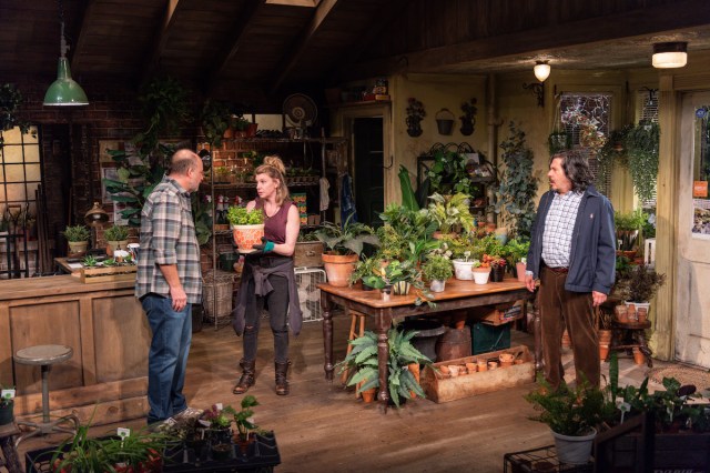 A man looks at a woman holding a potted plant. Another man with longer hair looks on aghast from the side. 