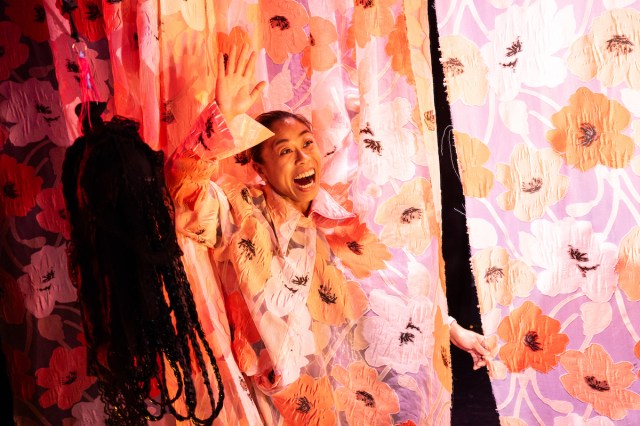 A woman waves and smile from within a curtain. 