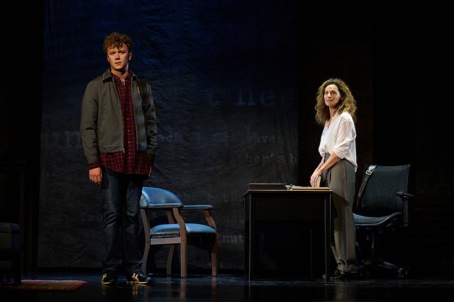 A man stads downstage of a woman behind a desk. 