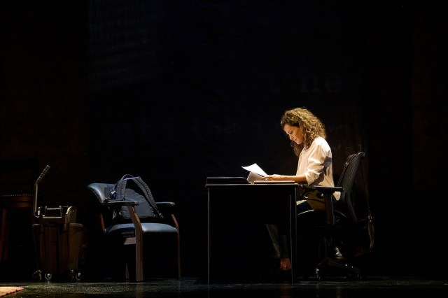 A woman sits at a desk on a dark stage and looks at papers. 