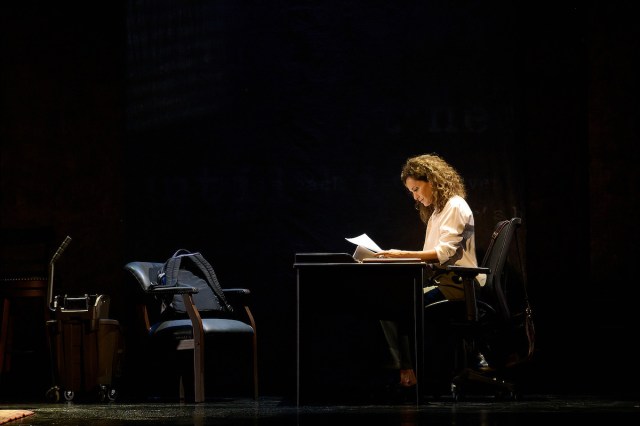 TSI 2 A woman sits at a desk on a dark stage and looks at papers.