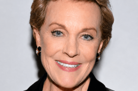 Smiling woman with short blonde hair, blue eyes, wearing a black blazer and small hoop earrings.